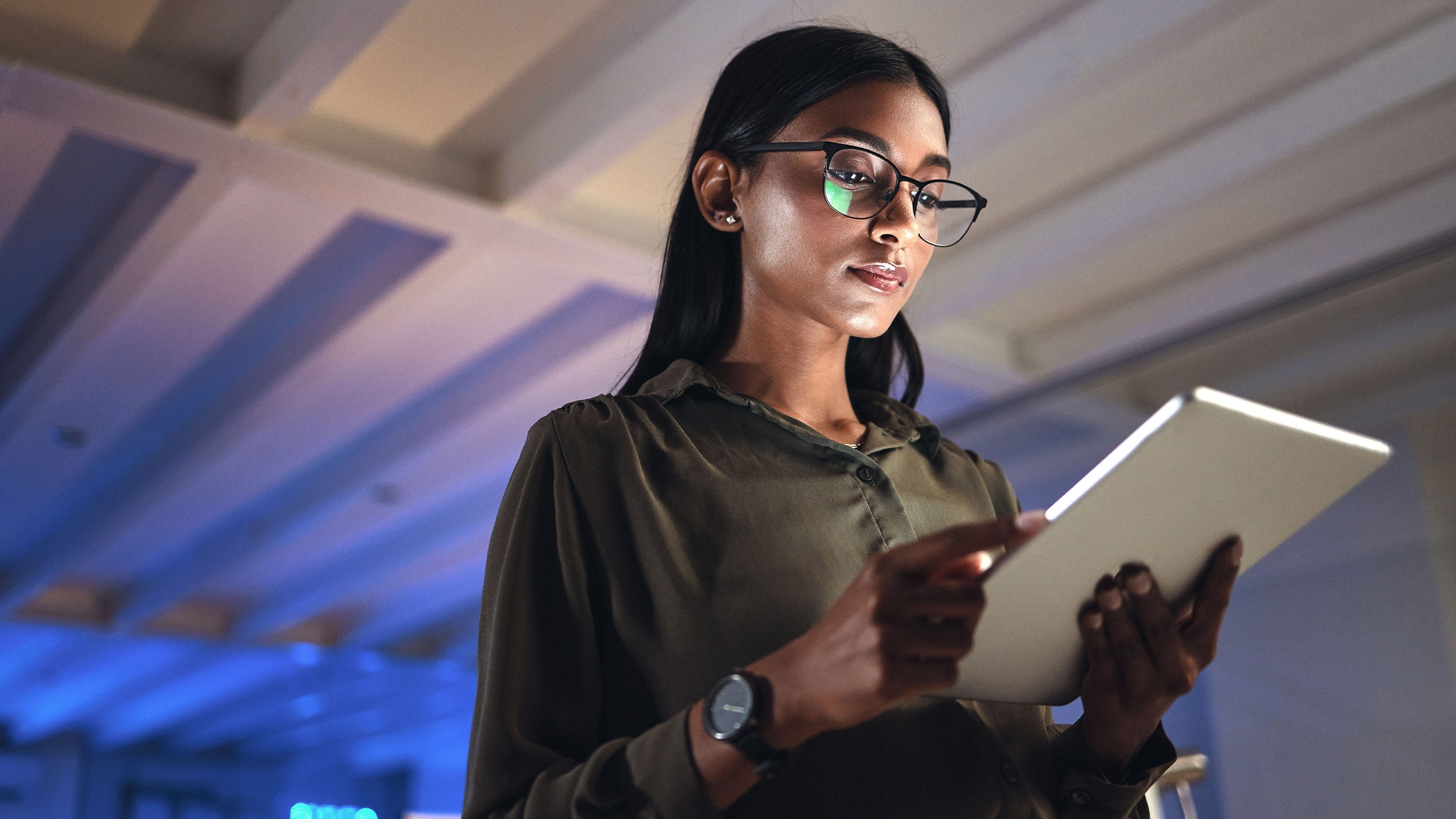 Young woman in glasses focuses on a digital tablet, illuminated by soft blue and white lighting, embodying modern tech and professional concentration.