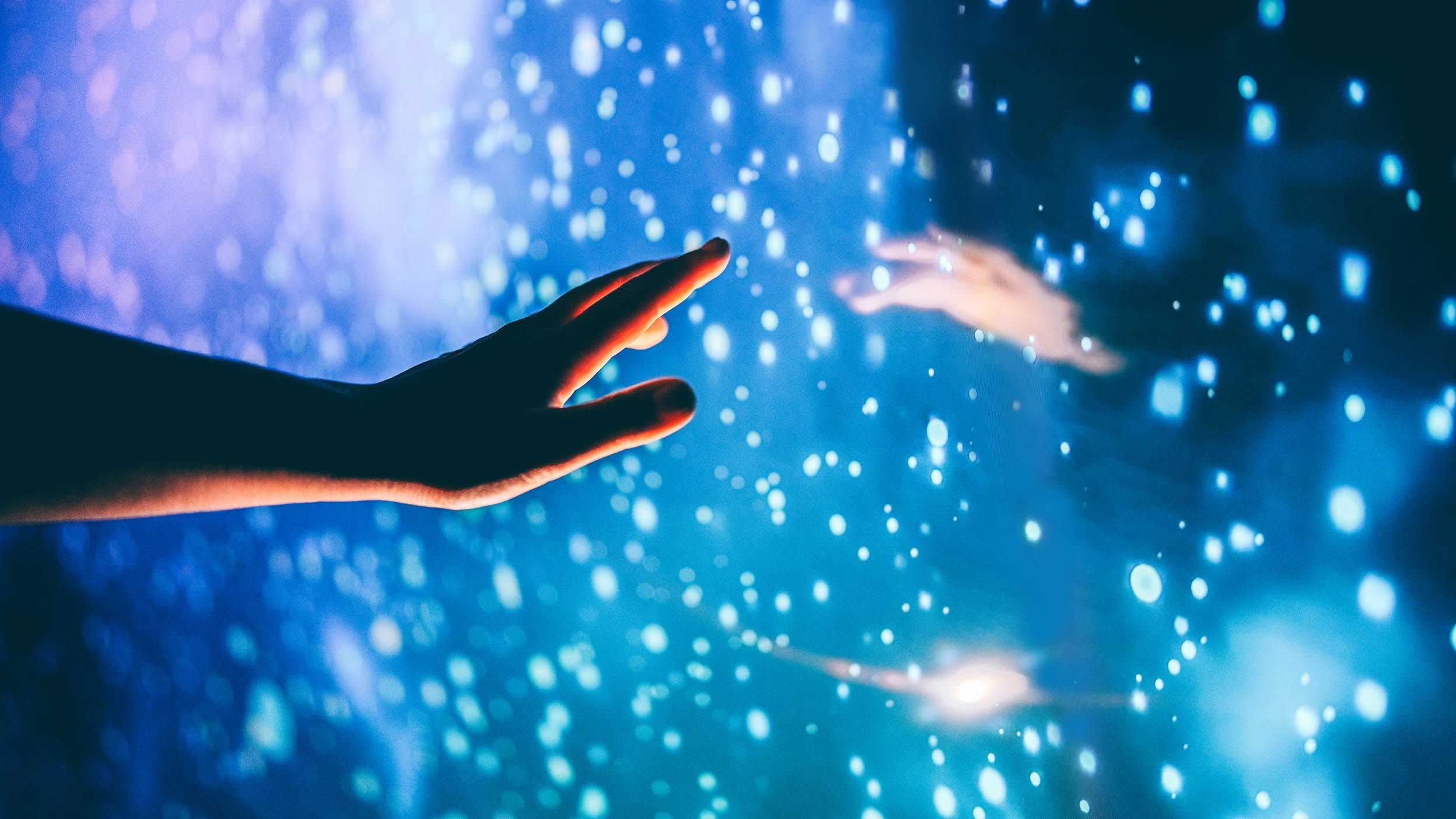 A hand reaches toward glowing blue particles on a cosmic backdrop—evoking touch, technology, and immersive interactivity.