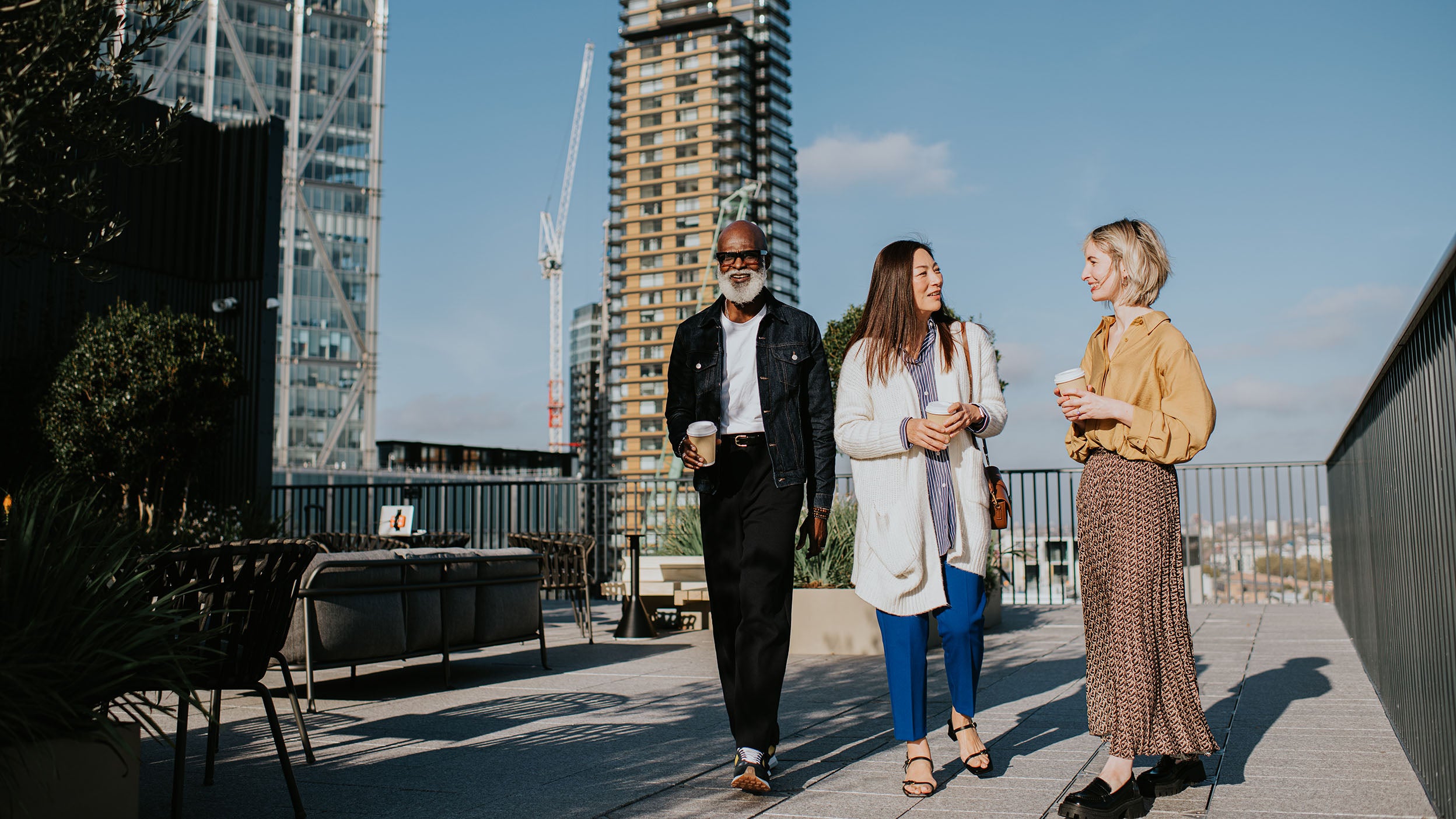 Three people in smart-casual business attire walk together on a roof top, surrounded by high rise buildings.