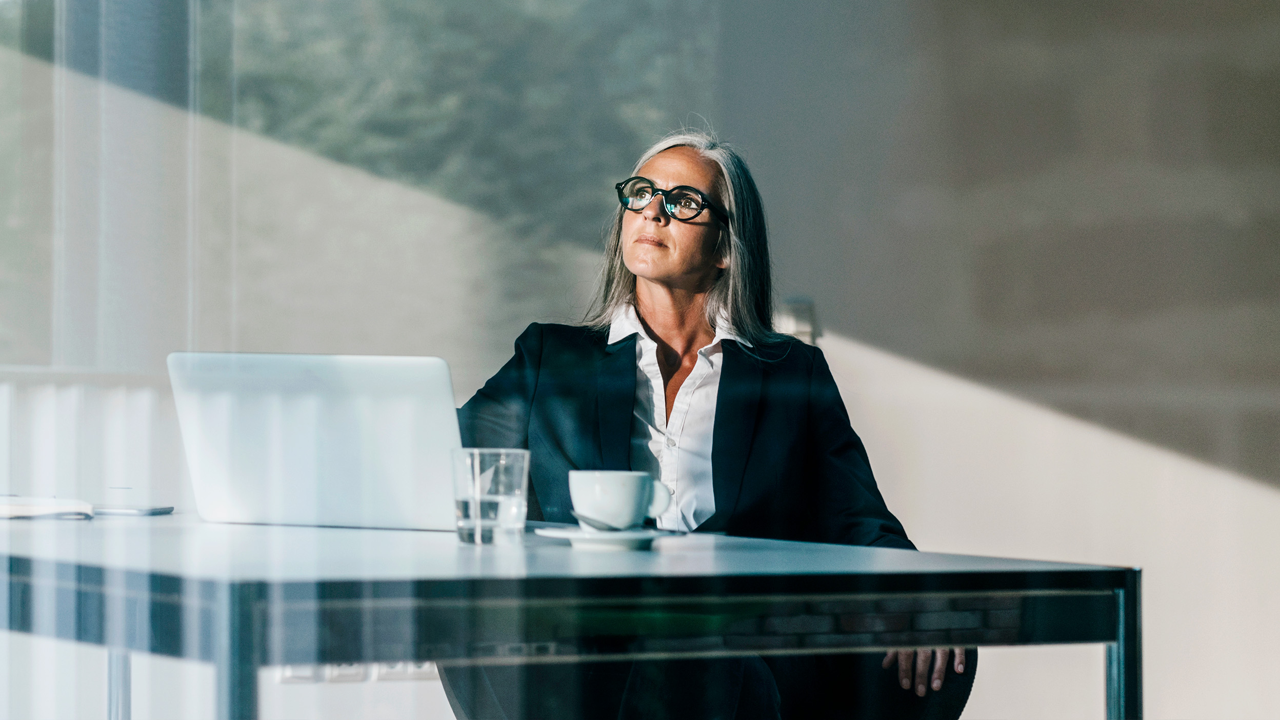 Woman sitting at desk with a cup and laptop, staring into the distance