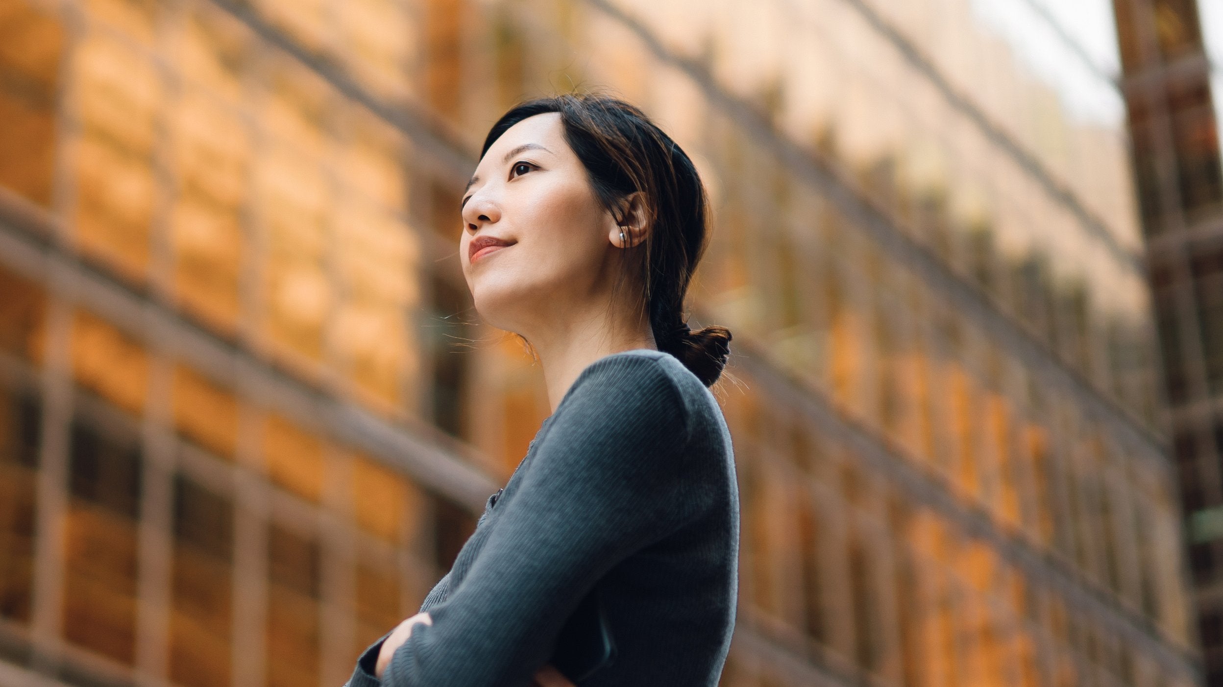 Person standing outdoors with arms crossed, wearing a long-sleeved top, in front of a modern glass building reflecting warm golden tones.