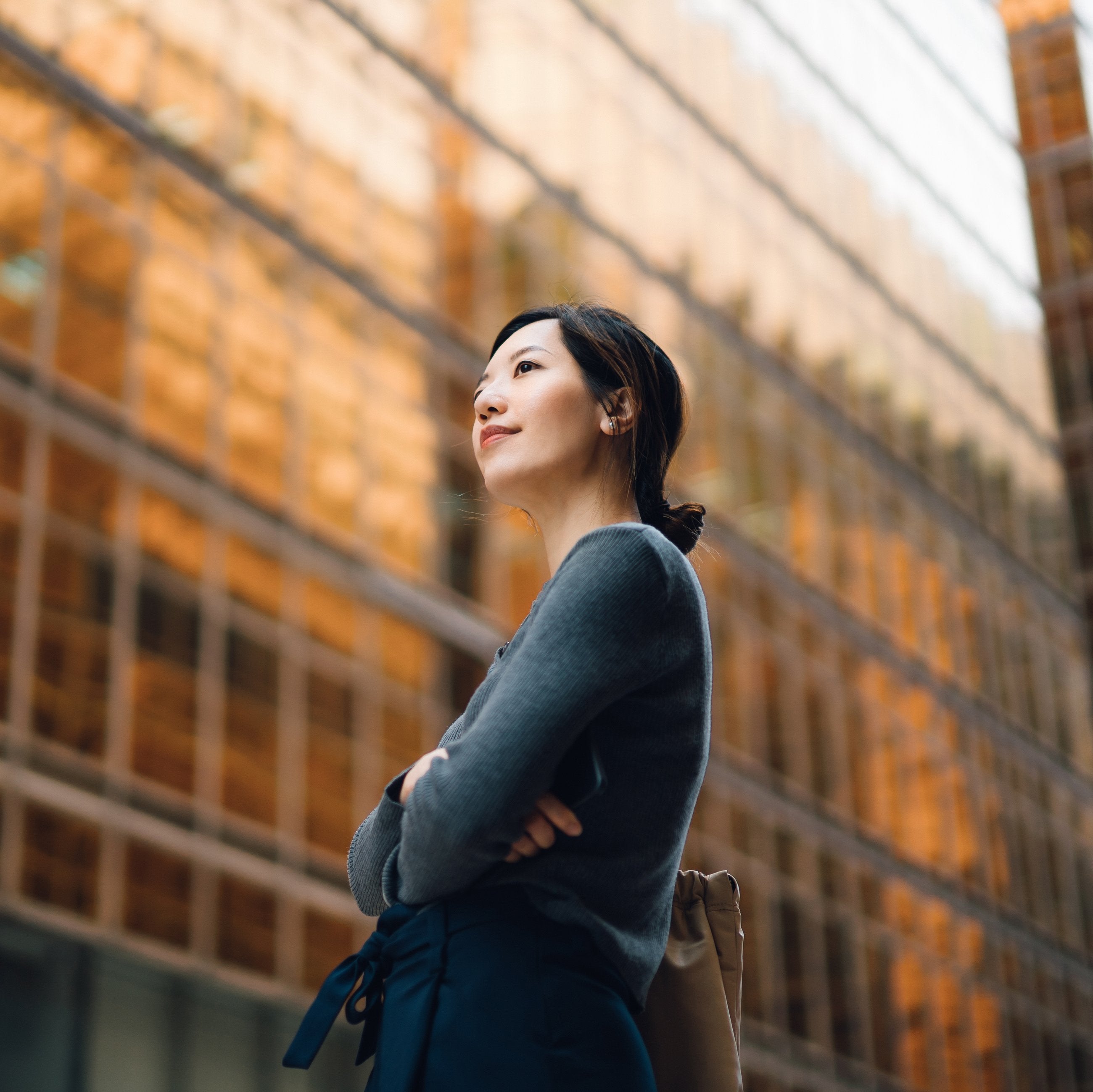 Person%20standing%20outdoors%20with%20arms%20crossed,%20wearing%20a%20long-sleeved%20top,%20in%20front%20of%20a%20modern%20glass%20building%20reflecting%20warm%20golden%20tones.