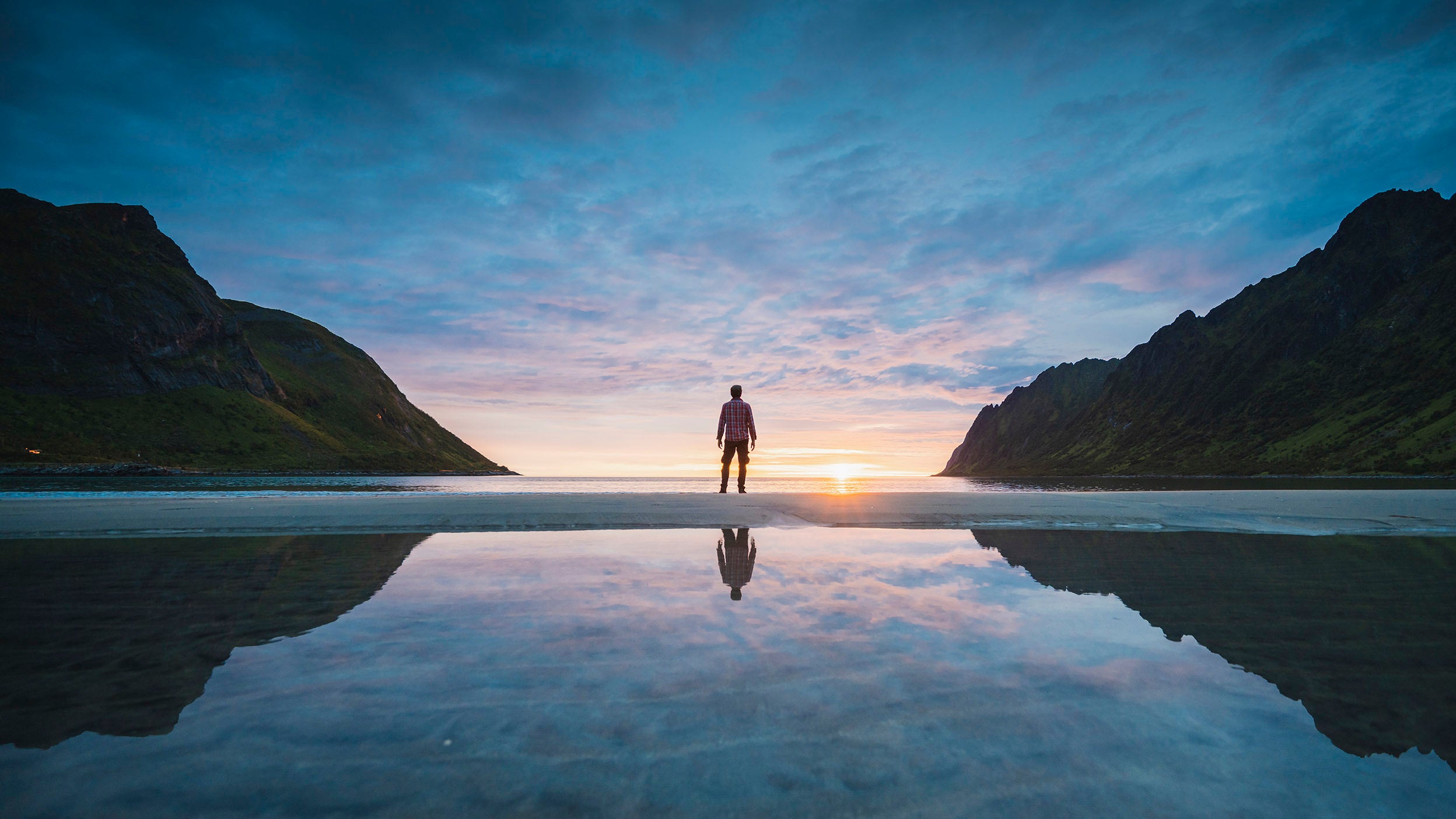 Person standing on a beach at sunrise, reflected in calm water between mountains.