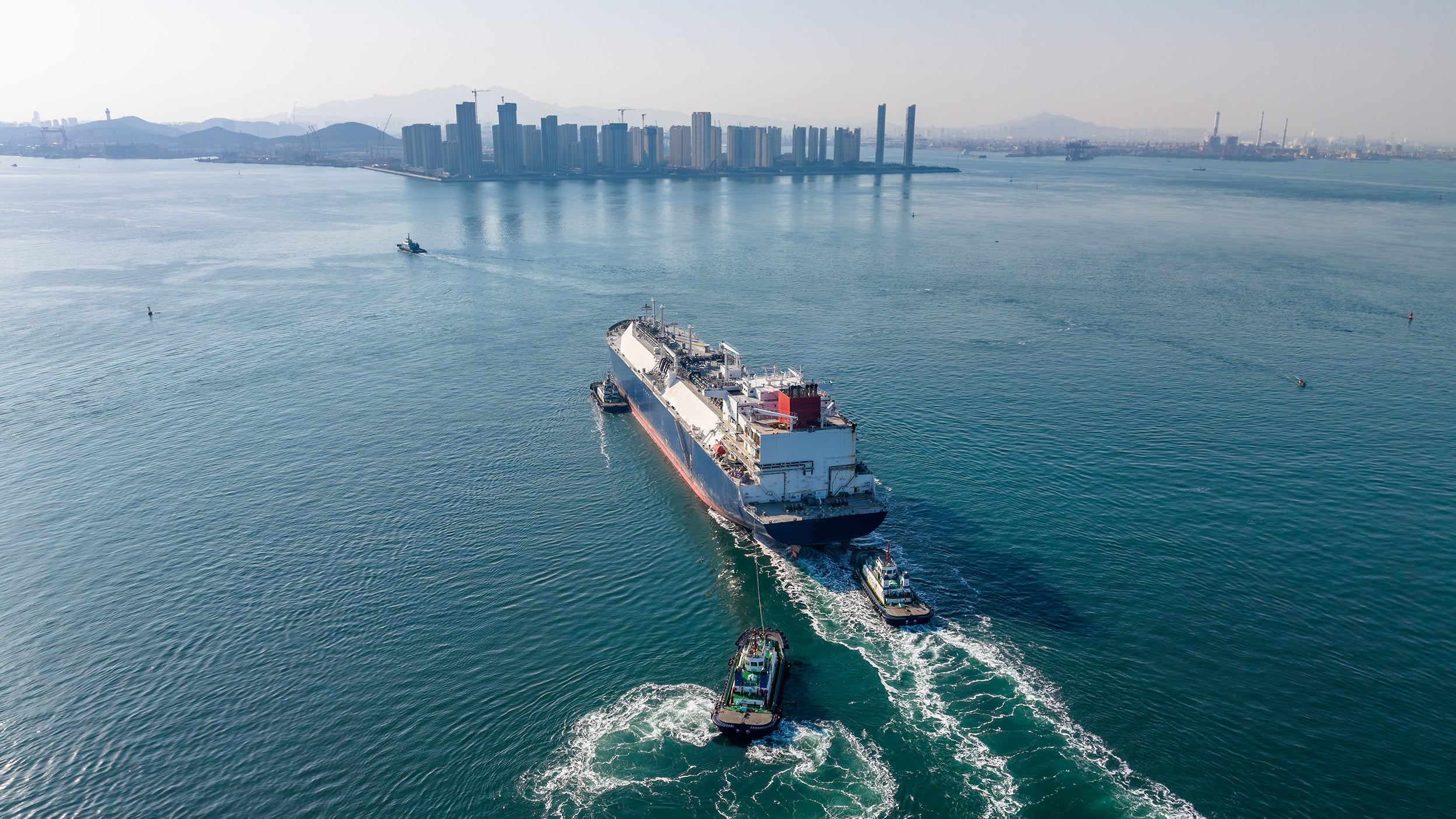 Cargo ship with tugboats near city and mountains.