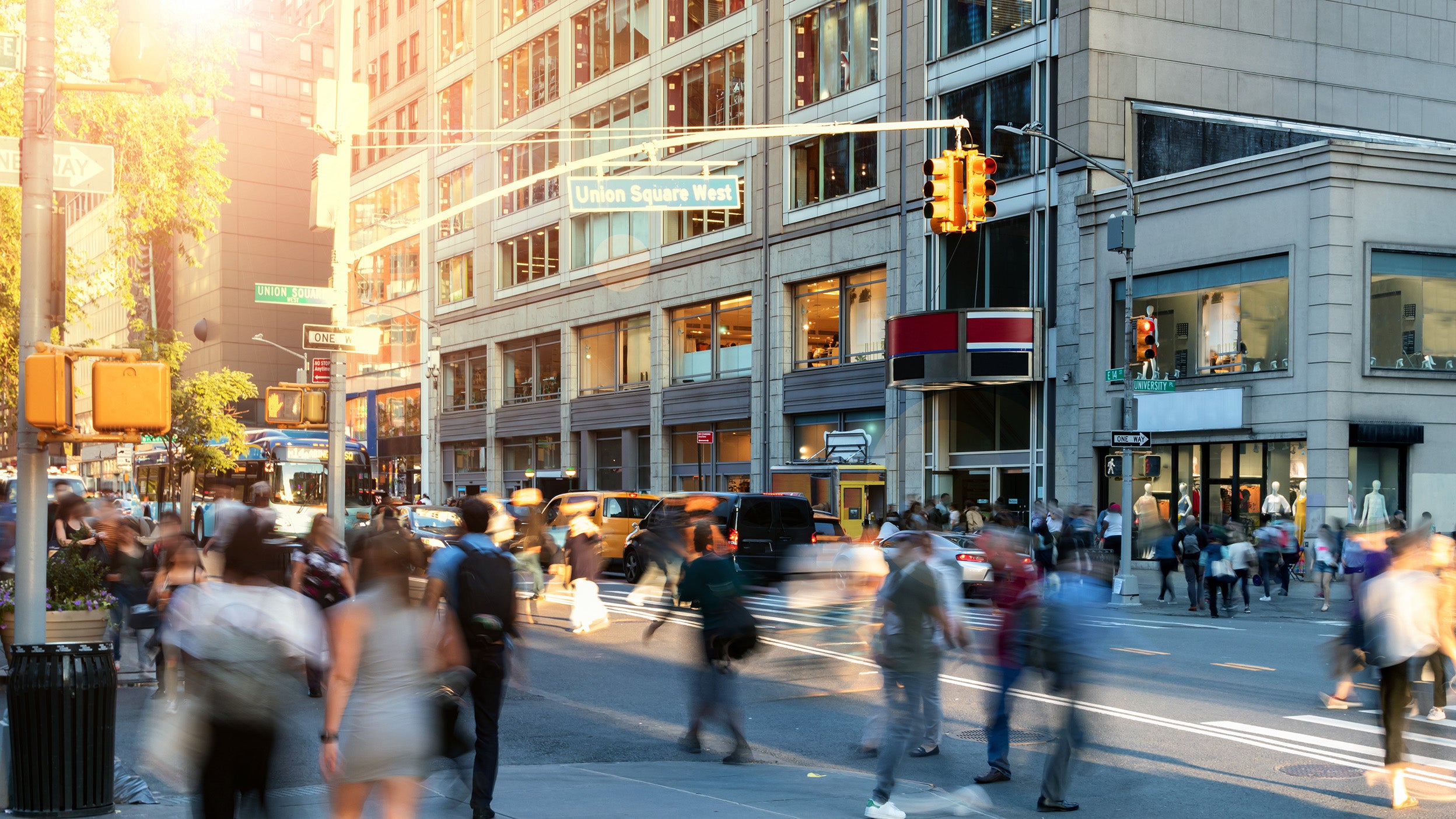 Crowds of people walking through the busy intersection on 14th Street at Union Square Park in New York City with sunlight shining in the background.