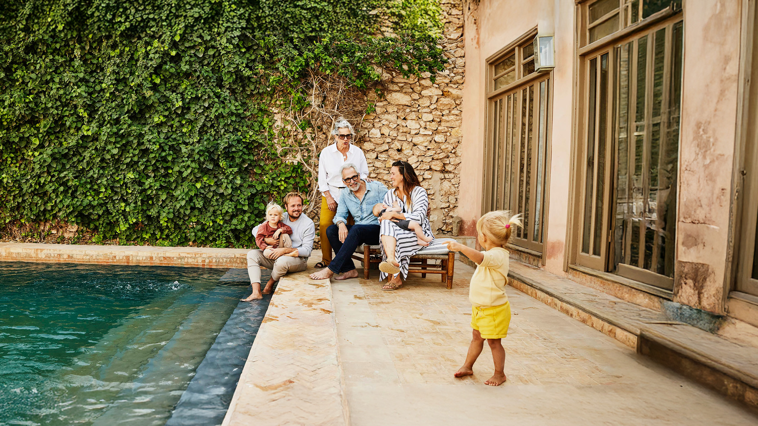 Wide shot of smiling grandparents relaxing with children and grandchildren beside pool at tropical villa during multigenerational family vacation