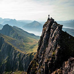 A%20person%20stands%20on%20top%20of%20a%20mountain%20looking%20at%20other%20mountain%20peaks.%20