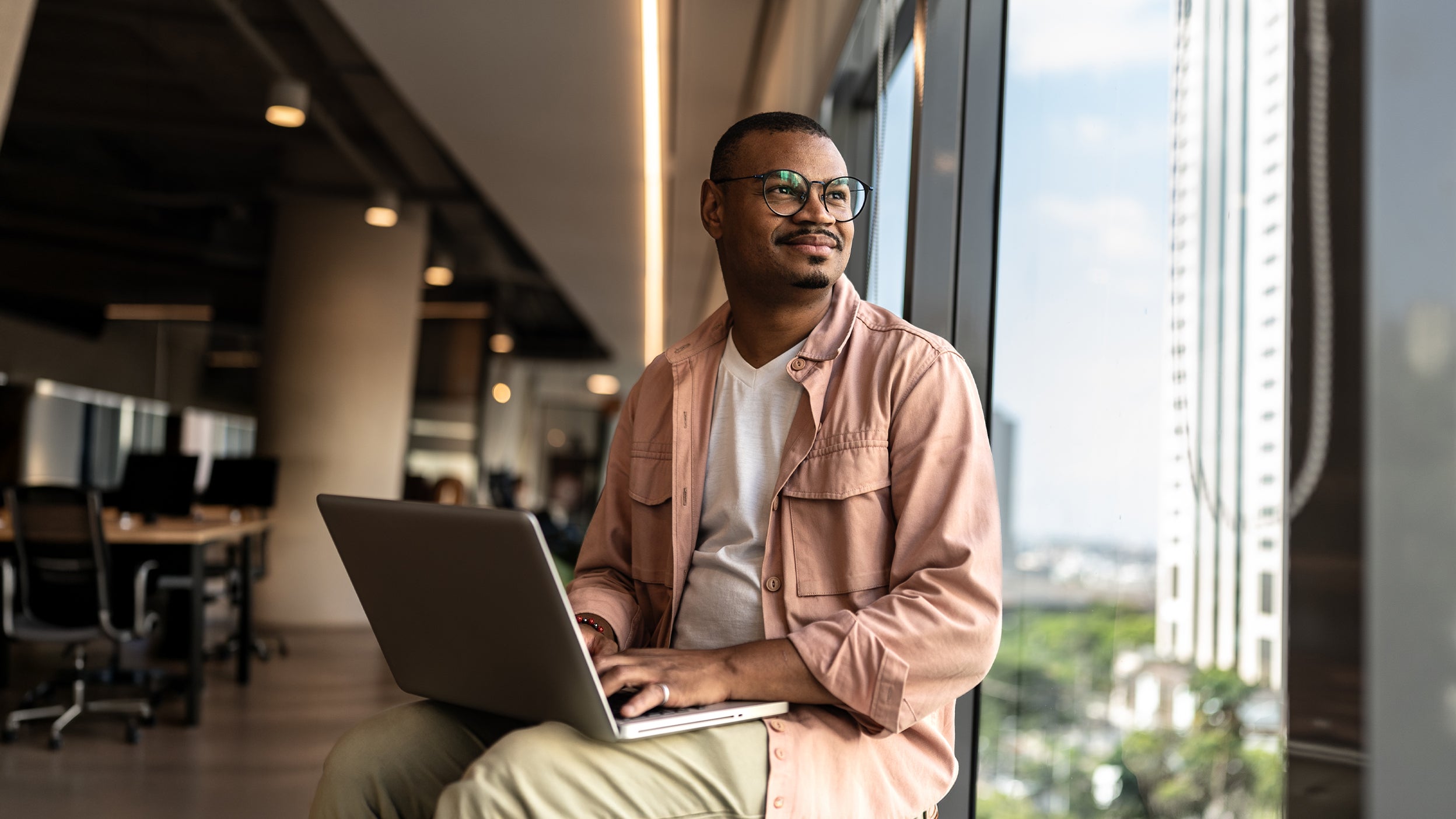Person by window with laptop on lap in modern indoor setting, seated comfortably.