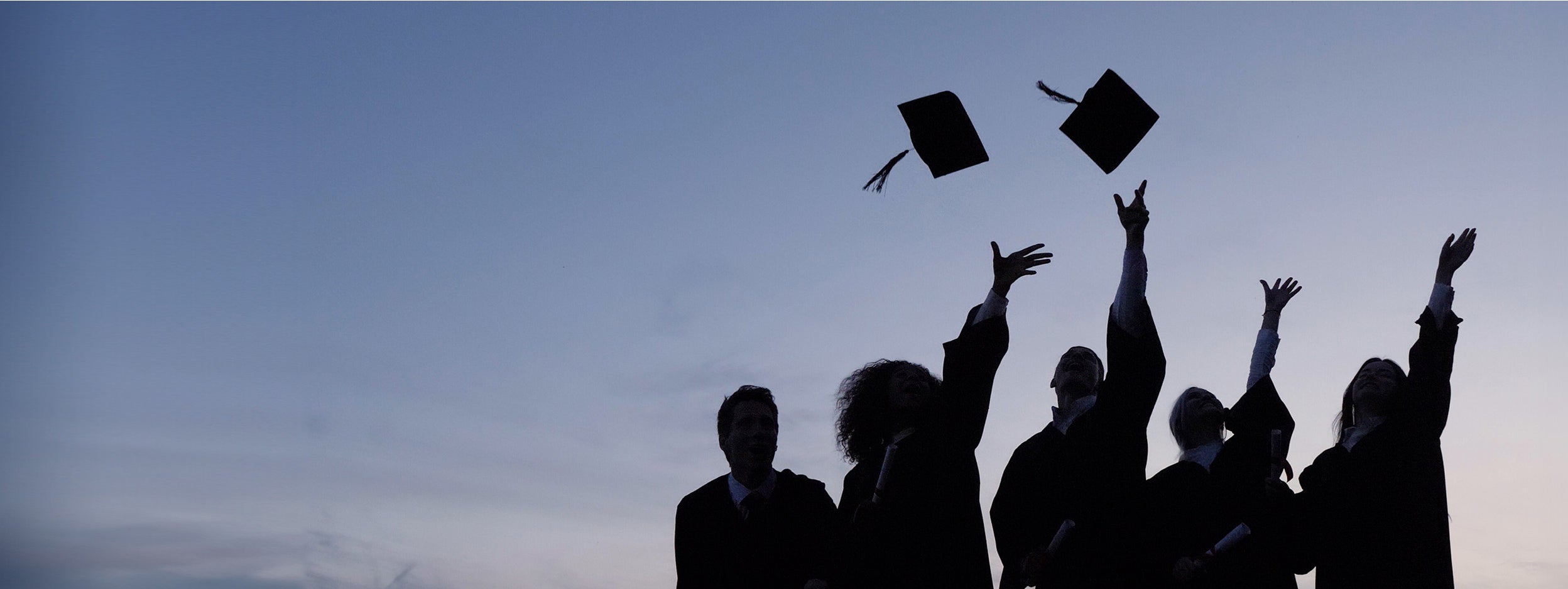 Silhouetted graduates throw caps into the air against a twilight sky